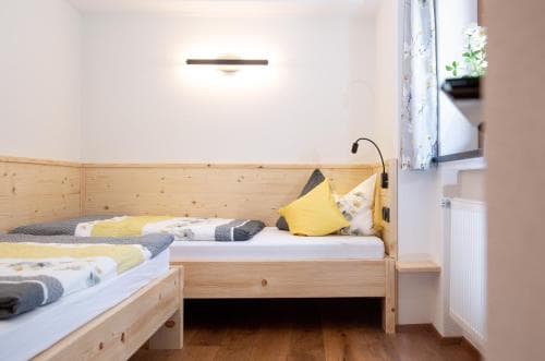 A cozy, minimalist bedroom with wooden bunk beds, soft yellow bedding, and natural light filtering through a window with floral curtains, ideal for travelers seeking a serene and comfortable stay in a budget-friendly accommodation.
