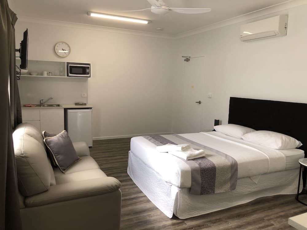 A cozy hotel room features a neatly made bed, a comfortable armchair, a small kitchenette with a microwave and sink, and a minimalist design, ideal for travelers seeking a clean and functional stay.
