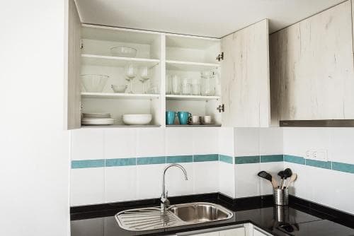A modern kitchen with open shelving displaying neatly arranged dishes, white tiles accented by teal stripes, and a sleek stainless steel sink creates a clean and inviting atmosphere ideal for travelers seeking a cozy self-catering accommodation.