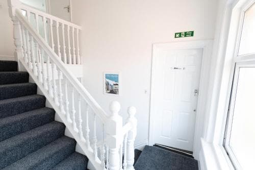 A bright and inviting hotel hallway features a white staircase with blue carpeted steps, a framed poster on the wall, and a door marked with an exit sign, ideal for travelers seeking a clean and accessible accommodation.