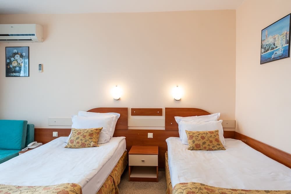 A cozy hotel room features two neatly made beds with floral pillows, warm wall-mounted lights, and artwork, creating a welcoming atmosphere ideal for travelers seeking comfort and relaxation.