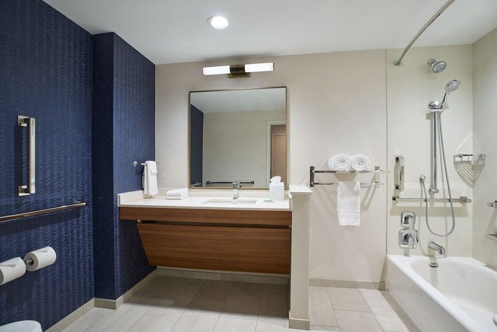 A modern hotel bathroom features a sleek wooden vanity, a bathtub with a handheld shower, neatly folded towels, and a calming blue accent wall, ideal for travelers seeking comfort and style during their stay.