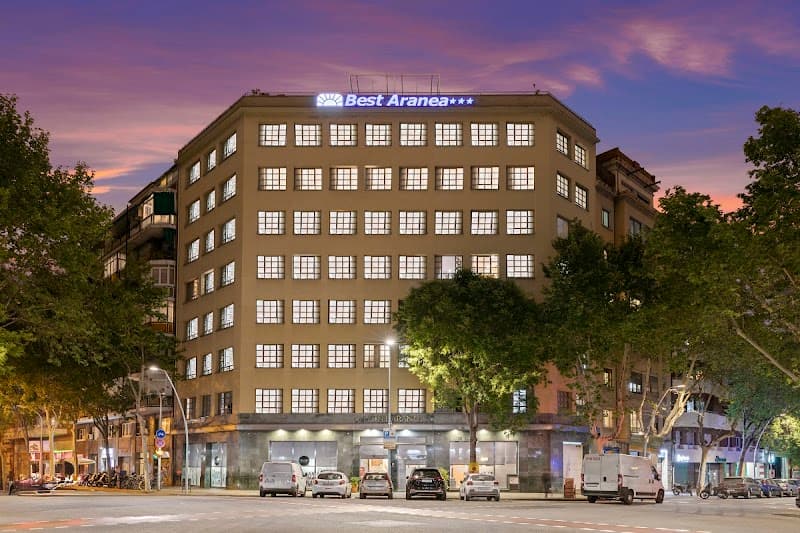 The Hotel Best Aranea in Barcelona's Eixample district glows warmly under the twilight sky, surrounded by lush trees and bustling streets, offering travelers a cozy urban retreat with easy access to city attractions.