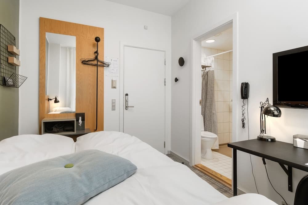 A cozy hotel room features a neatly made bed, a desk with a lamp, a mirror with hangers, and an open bathroom door, creating a clean and functional space ideal for travelers seeking comfort and convenience.