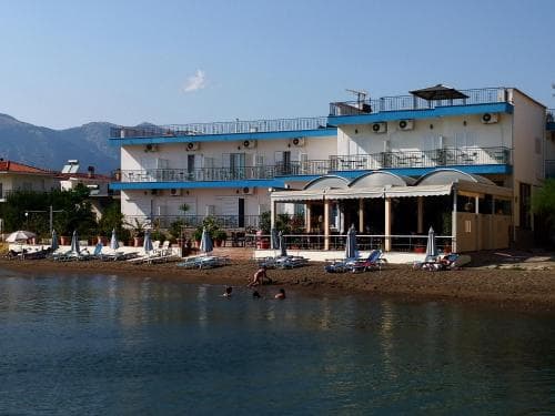 A serene coastal scene features a beachside hotel with blue balconies, surrounded by lounge chairs and umbrellas under clear skies, ideal for travelers seeking relaxation near the water.