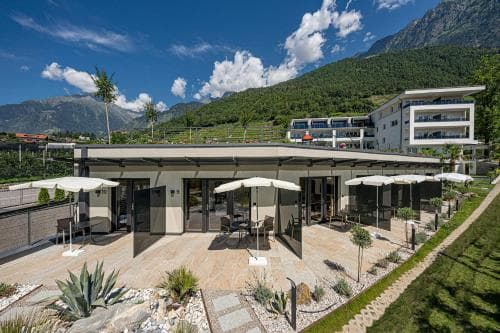 A serene mountain retreat features a modern patio with white umbrellas and lounge chairs, set against lush greenery and towering peaks, ideal for travelers seeking relaxation and scenic views.