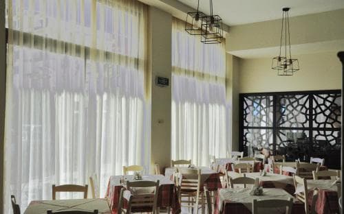 A cozy indoor dining area bathed in soft natural light from large windows, featuring neatly arranged tables with red accents, modern hanging lamps, and a decorative lattice wall, ideal for travelers seeking a relaxed meal spot.
