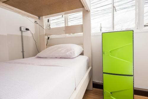 A cozy hostel room features a neatly made bed, bright lime-green storage drawers, and large windows letting in natural light, ideal for budget travelers seeking comfort and simplicity.