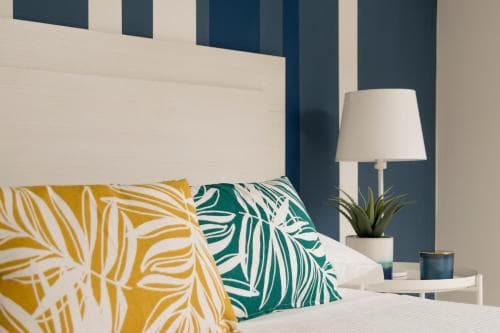 A cozy bedroom scene features a bed adorned with vibrant tropical-patterned pillows against a stylish navy-striped wall, complemented by a modern lamp and potted plant, ideal for travelers seeking a relaxing and aesthetically pleasing accommodation.