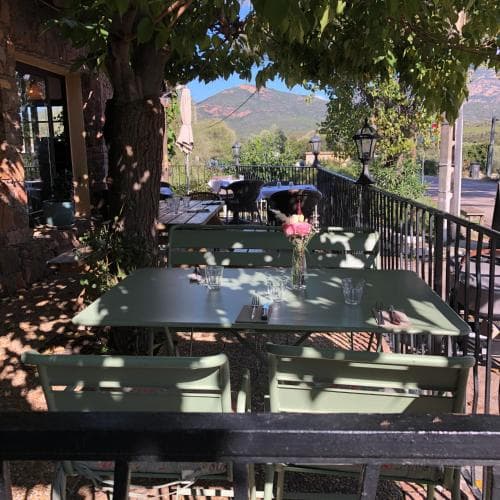 A serene outdoor café scene with green tables and chairs under leafy trees, set against a picturesque mountain backdrop, invites travelers to relax and enjoy a leisurely meal in a tranquil, rustic setting.