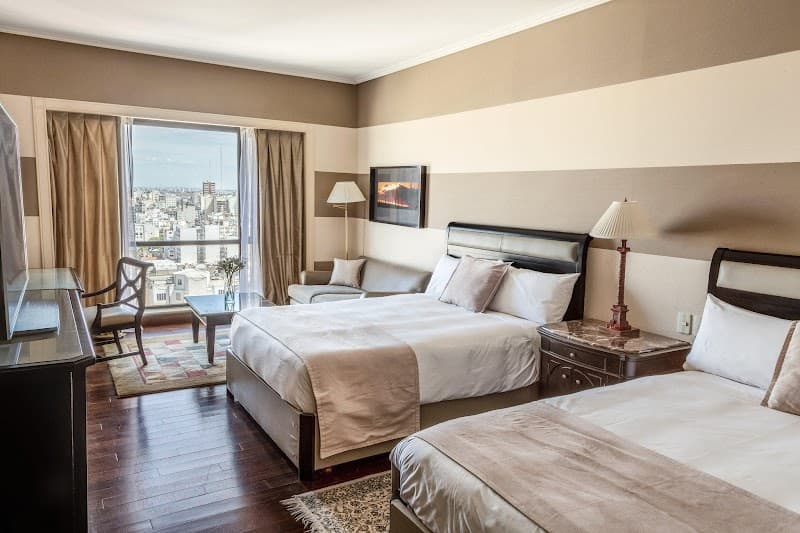 A cozy hotel room at the InterContinental Buenos Aires by IHG features two neatly made beds, a comfortable seating area, and a stunning city view through large windows, ideal for travelers seeking relaxation and urban exploration.