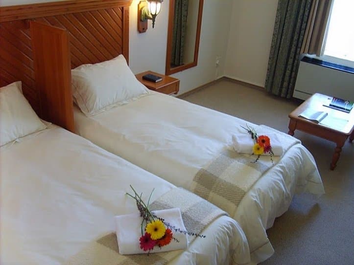A cozy hotel room features two neatly made beds adorned with fresh flowers, a wooden nightstand with a lamp, and a small table by the window, ideal for a comfortable stay or romantic getaway.