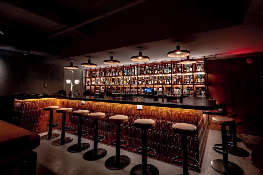 A cozy, dimly-lit bar with warm pendant lights, patterned walls, and an extensive liquor display invites travelers seeking a sophisticated nightlife experience.