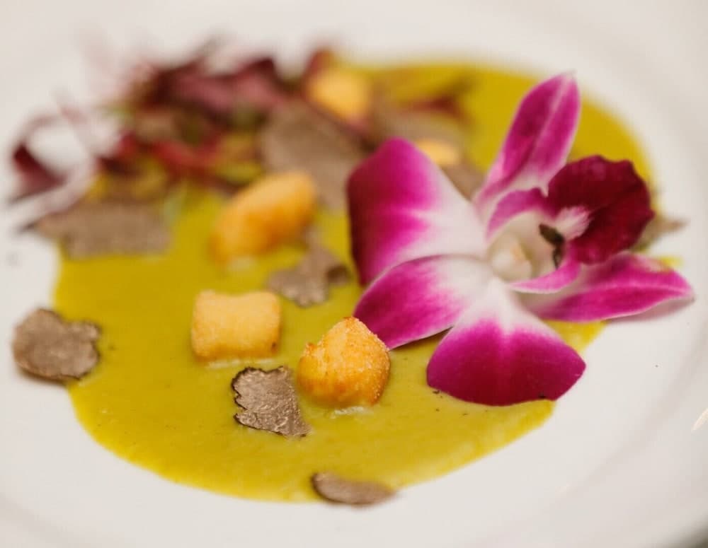 A gourmet dish featuring a vibrant yellow sauce, crispy croutons, delicate truffle slices, and a striking purple orchid garnish creates an elegant fine dining experience perfect for culinary enthusiasts exploring high-end restaurants.