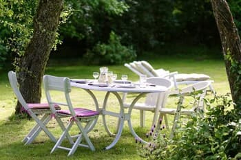 A serene outdoor dining setup with white chairs and a table surrounded by lush greenery, perfect for a tranquil garden retreat or a relaxing picnic experience.