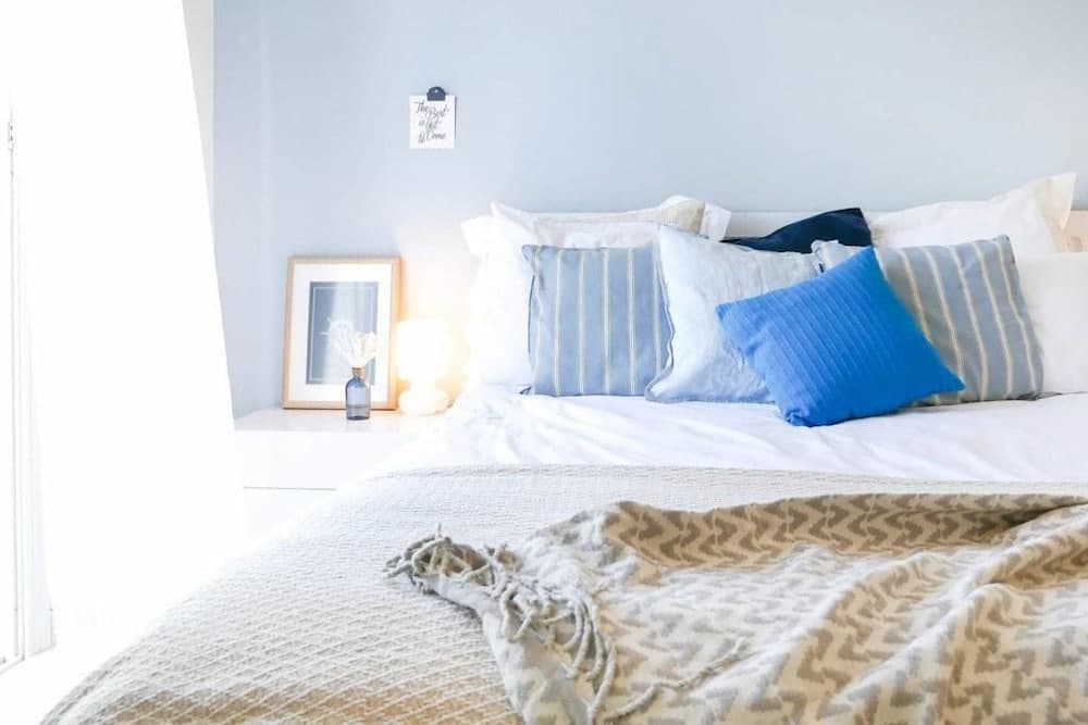 A serene bedroom with soft blue walls, cozy bedding adorned with blue pillows and a patterned throw, a framed picture, and a glowing lamp creates an inviting retreat ideal for relaxation and travel accommodation inspiration.