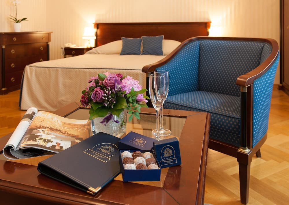 A cozy hotel room exudes warmth with its inviting bed, plush blue armchair, and a coffee table adorned with chocolates, flowers, and a magazine, perfect for a relaxing getaway.