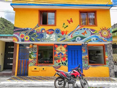 A vibrant yellow building adorned with colorful mosaic art featuring butterflies and floral patterns stands in a lively street scene, perfect for travelers seeking unique accommodations and cultural experiences.