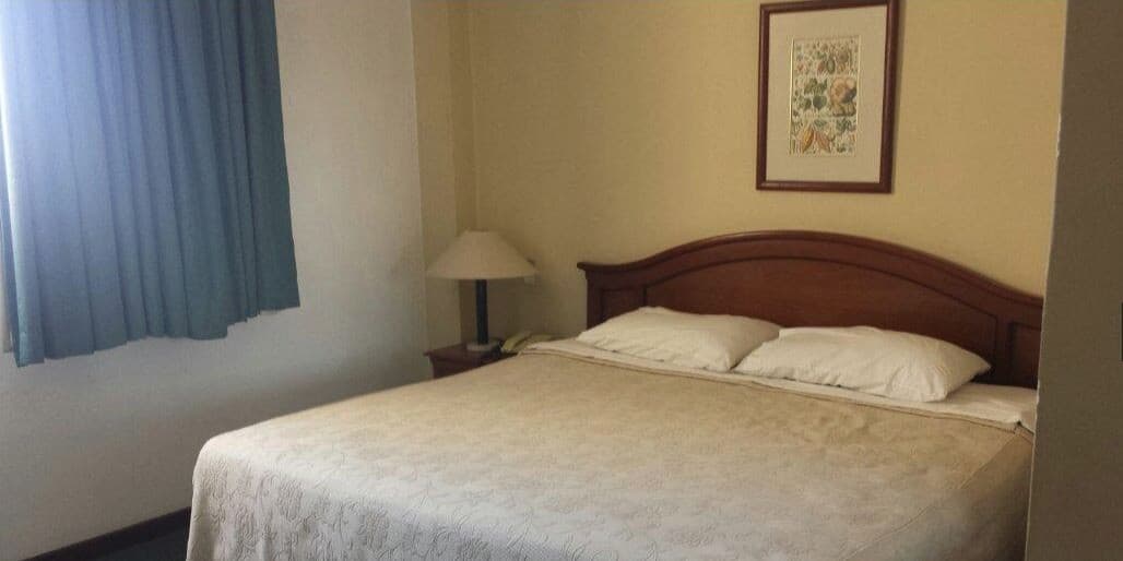 A cozy hotel room features a neatly made bed with white linens, a wooden headboard, a bedside lamp, and a framed picture on the wall, ideal for travelers seeking comfort and simplicity.