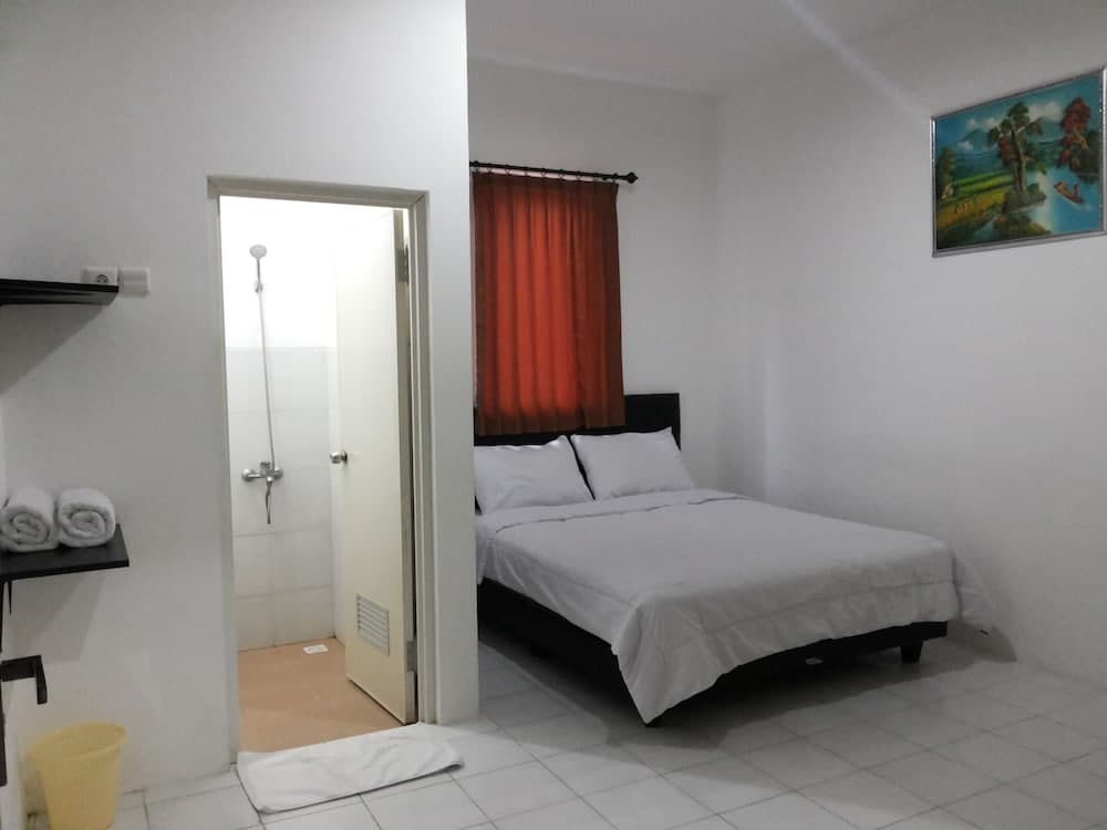 A cozy budget-friendly hotel room features a neatly made bed, vibrant red curtains, a small bathroom with a shower, and a serene landscape painting, ideal for travelers seeking simplicity and comfort.