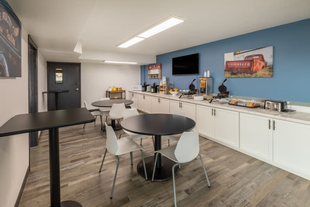 A cozy hotel breakfast area features modern black tables, white chairs, a blue accent wall adorned with vintage café artwork, and a well-equipped kitchenette, ideal for travelers seeking a comfortable morning start.