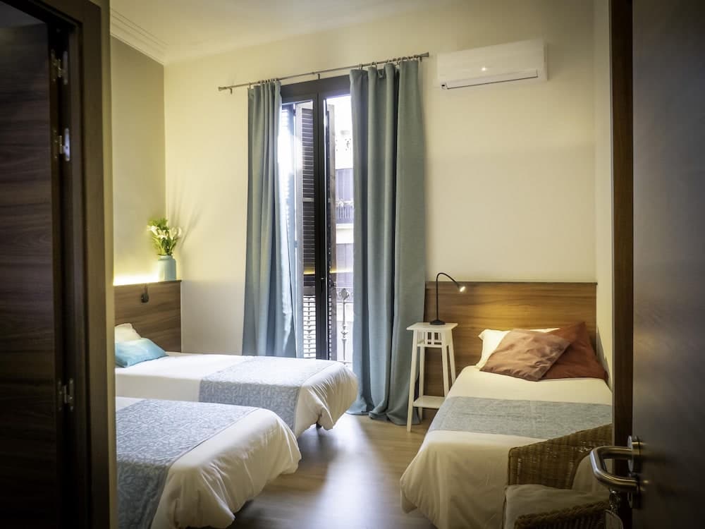 A cozy hotel room with neatly arranged twin beds, soft blue curtains framing a balcony view, and warm lighting creates an inviting atmosphere ideal for travelers seeking comfort and relaxation.