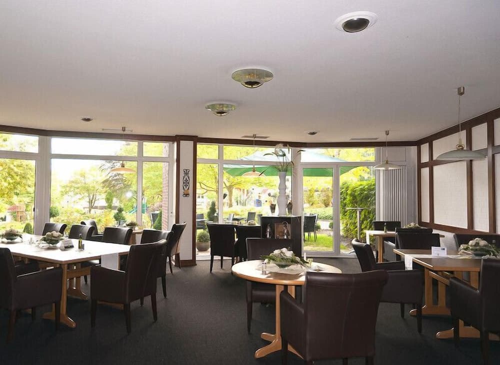 A cozy restaurant with large windows overlooking lush greenery offers a serene dining experience with neatly arranged tables, chairs, and floral centerpieces, ideal for travelers seeking a peaceful meal in a natural setting.