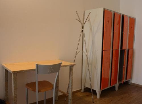 A cozy hostel room features a simple wooden desk with a chair, a minimalist coat rack, and vibrant orange lockers against a neutral wall, ideal for budget travelers seeking functional and compact accommodations.