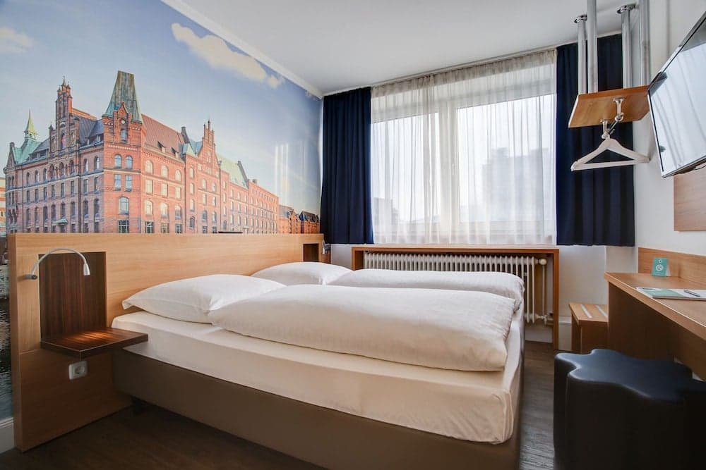 A cozy hotel room features a neatly made bed beside a large window with blue curtains, a desk area, and a vibrant cityscape mural, ideal for travelers seeking comfort and a touch of local charm.