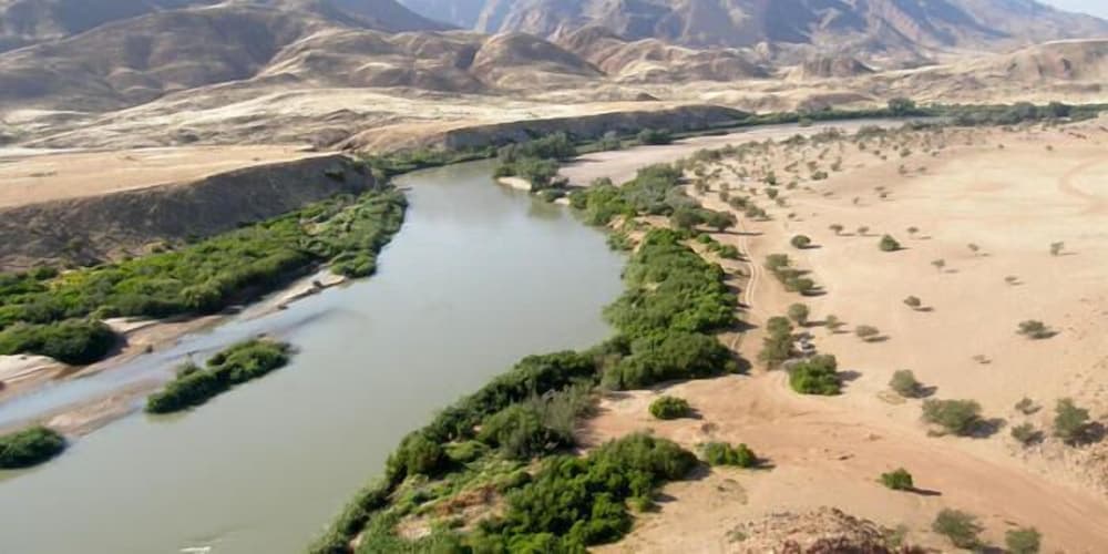 A serene river winds through a lush oasis amidst arid desert landscapes, offering travelers a tranquil retreat with scenic views of distant mountains and vibrant greenery ideal for nature photography and eco-tourism.
