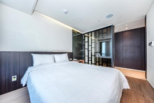 A modern hotel room features a neatly made bed with white linens, sleek wooden furnishings, and a glass-enclosed bathroom, creating a clean and minimalist atmosphere ideal for travelers seeking comfort and style.