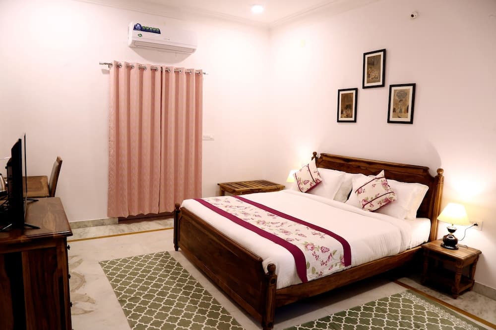 A cozy hotel room features a wooden bed adorned with floral bedding, soft pink curtains, and a desk with a TV, ideal for travelers seeking comfort and relaxation.