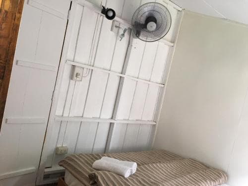 A cozy hostel room features a simple bed with striped bedding, a neatly folded towel, white wooden paneling, and a wall-mounted fan, ideal for budget travelers seeking comfort in a minimalist setting.