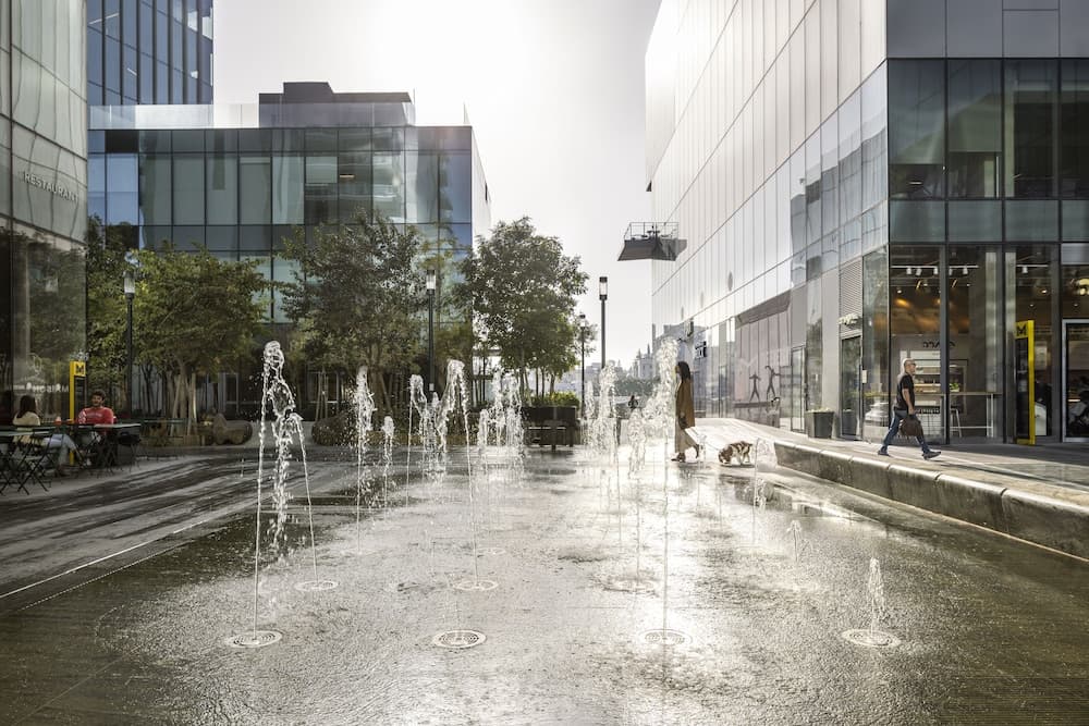 A vibrant urban plaza features playful water fountains surrounded by modern glass buildings, lush greenery, and pedestrians enjoying the sunny day, ideal for city exploration and relaxation.