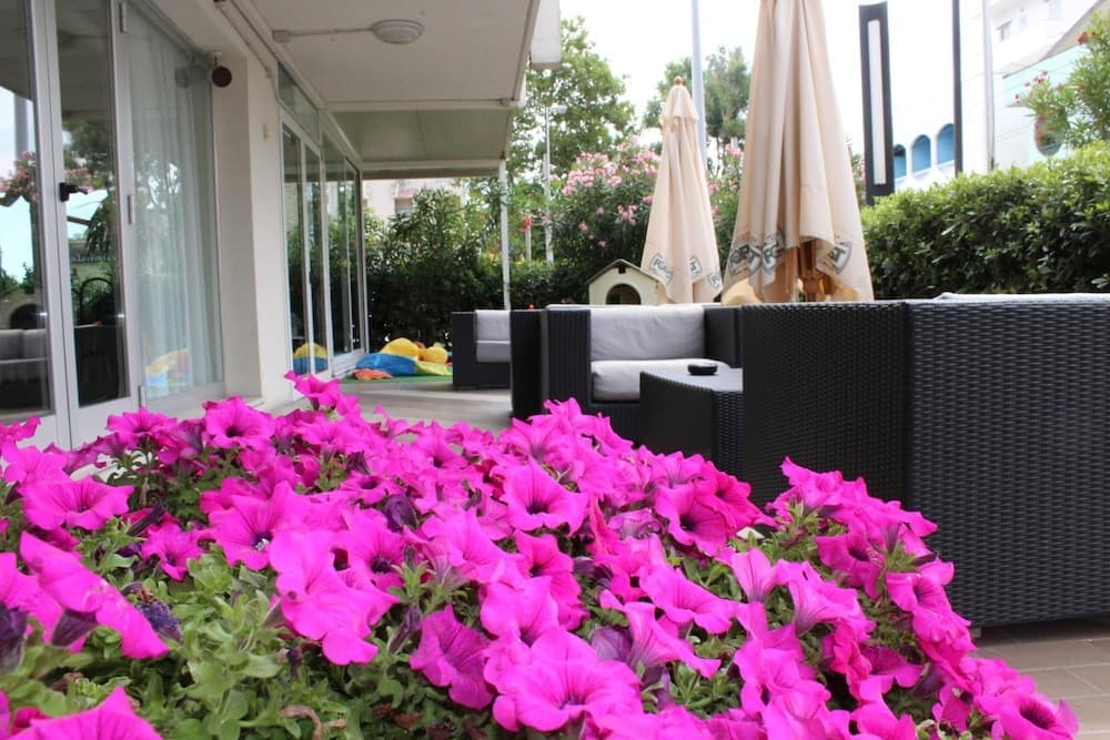 A serene outdoor patio features vibrant pink flowers in the foreground, cozy wicker furniture, and closed beige umbrellas, creating an inviting atmosphere perfect for relaxation or a romantic getaway.