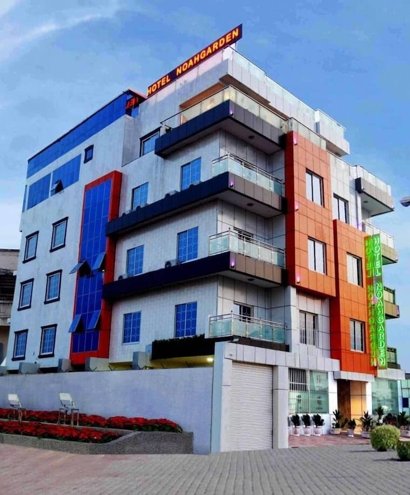 A vibrant, modern hotel with colorful panels and balconies stands invitingly against a clear sky, featuring potted plants and flower beds that enhance its welcoming atmosphere, ideal for travelers seeking a stylish urban stay.