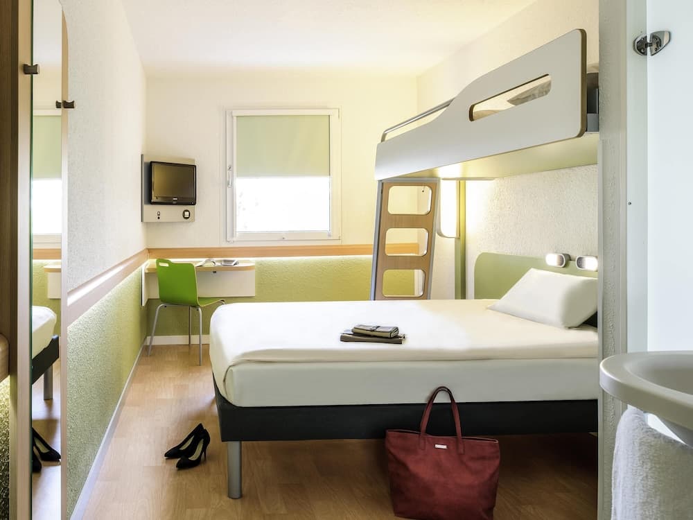 A cozy hotel room with a bunk bed setup, green accents, a small desk by the window, and personal items like shoes and a bag, ideal for budget travelers seeking comfort and functionality.