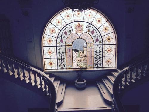 A serene indoor staircase scene features an ornate stained-glass window depicting a cityscape, framed by elegant railings, ideal for cultural heritage tourism photography.