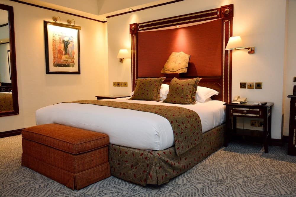 A cozy hotel room features a neatly made bed with patterned bedding, warm lighting from wall-mounted lamps, and a decorative ottoman, ideal for travelers seeking comfort and relaxation.