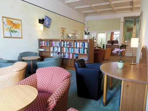A cozy hotel lounge features comfortable seating, a bookshelf filled with reading materials, and warm decor, creating an inviting atmosphere ideal for travelers seeking relaxation or a quiet spot to unwind.