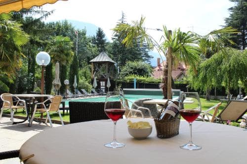 A serene outdoor patio features glasses of red wine on a table surrounded by lush greenery, palm trees, and a pool, ideal for a relaxing vacation retreat.