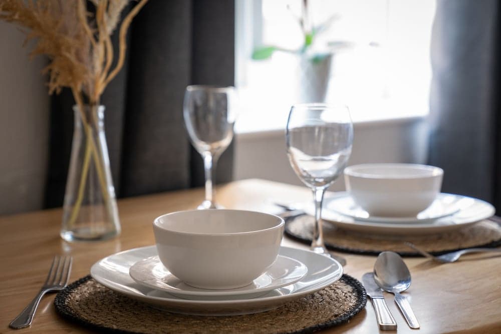 A cozy dining setup with white bowls, plates, wine glasses, and natural decor on a wooden table by a window, ideal for intimate gatherings or romantic dinners.