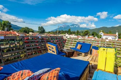 A vibrant rooftop lounge adorned with colorful wooden furniture and lush greenery offers panoramic views of distant mountains under a bright blue sky, perfect for travelers seeking relaxation and scenic beauty.