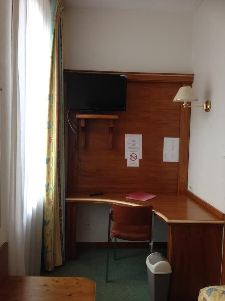 A cozy hotel room corner features a wooden desk with a chair, mounted TV, wall lamp, and floral curtains, ideal for travelers seeking a functional workspace in a serene setting.