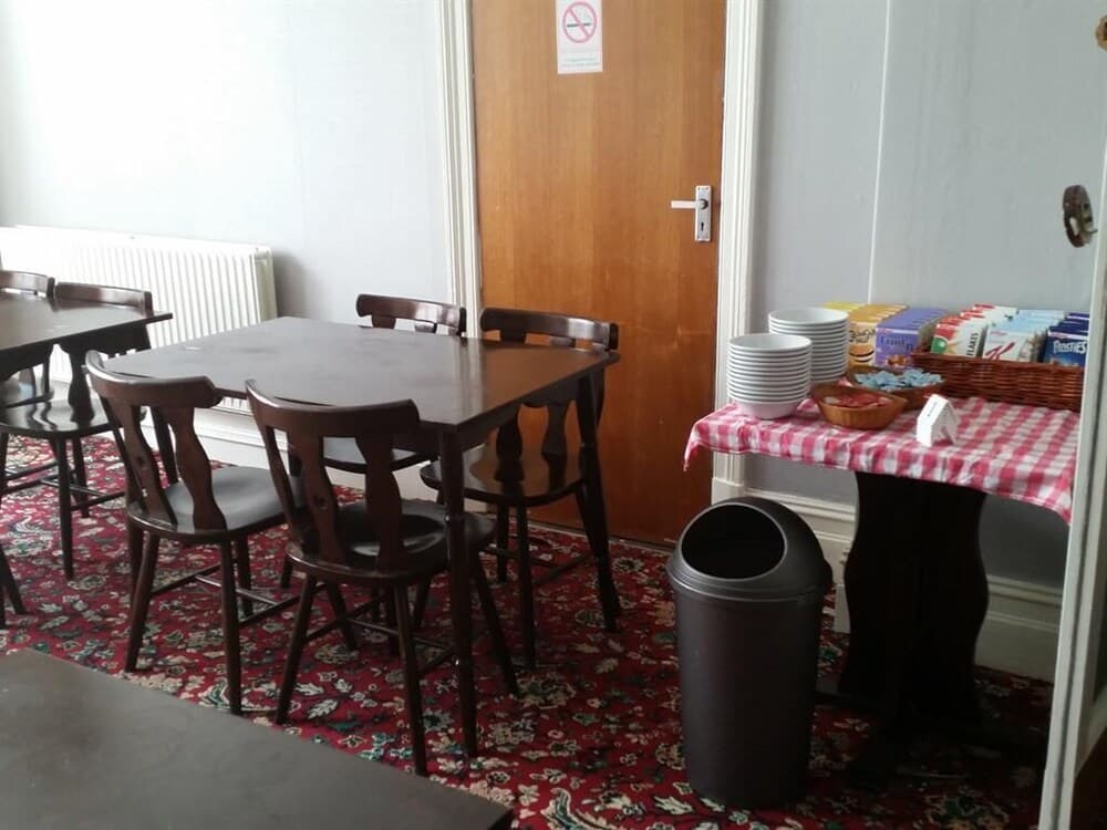 A cozy dining area with wooden tables and chairs on a patterned rug, featuring a checkered tablecloth with stacked plates and snacks, ideal for travelers seeking a quaint and welcoming breakfast spot.