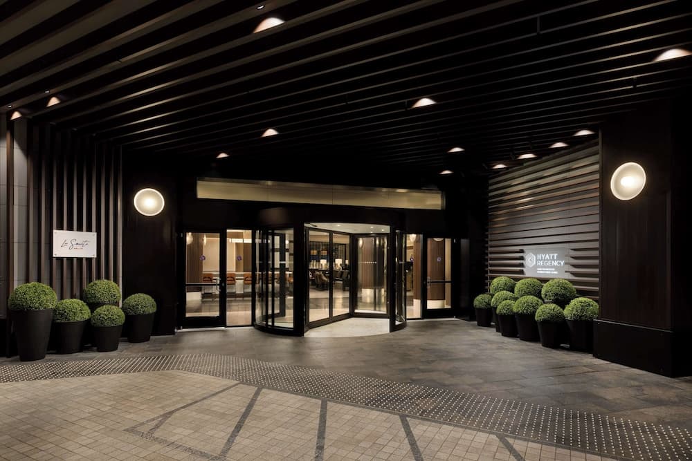 A modern hotel entrance featuring sleek black wood paneling, warm lighting, neatly trimmed shrubs in large pots, and glass doors leading into a well-lit interior, ideal for travelers seeking luxury accommodations.