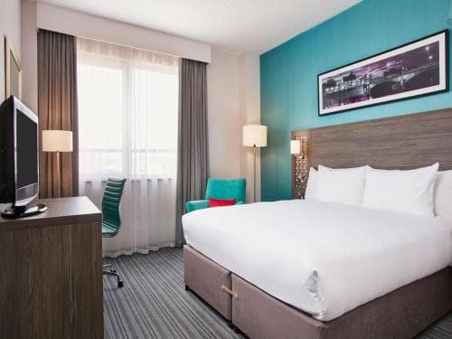 A cozy hotel room features a neatly made bed against a vibrant blue wall, modern furnishings including a desk and chair, and soft natural light filtering through sheer curtains, ideal for travelers seeking comfort and style during their stay.