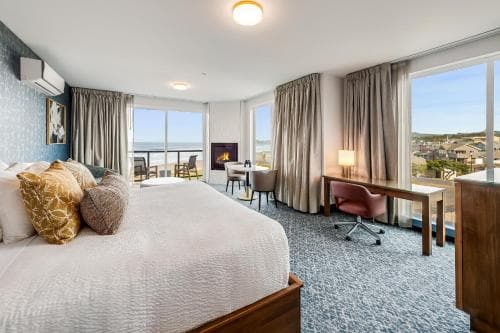 A cozy hotel suite with a plush bed, fireplace, and large windows overlooking the ocean offers a serene retreat for travelers seeking relaxation and comfort.