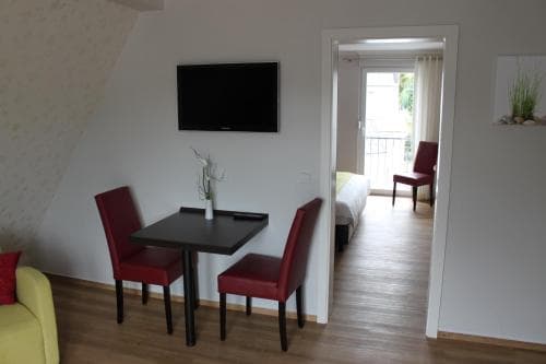 A cozy apartment interior features a small dining area with red chairs and a flat-screen TV, leading to a bright bedroom with a balcony, ideal for a comfortable city getaway.