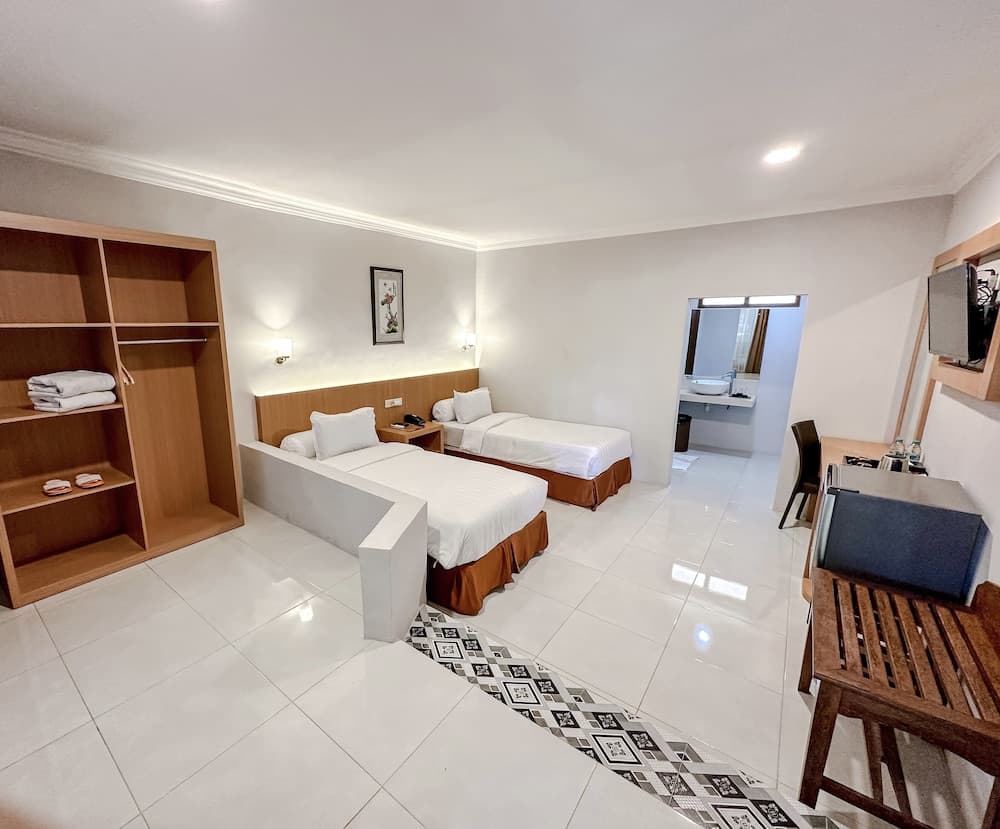 A cozy hotel room features two neatly made beds, a wooden shelving unit with folded towels, a small dining area, and a vanity mirror, creating a welcoming atmosphere ideal for travelers seeking comfort and convenience.